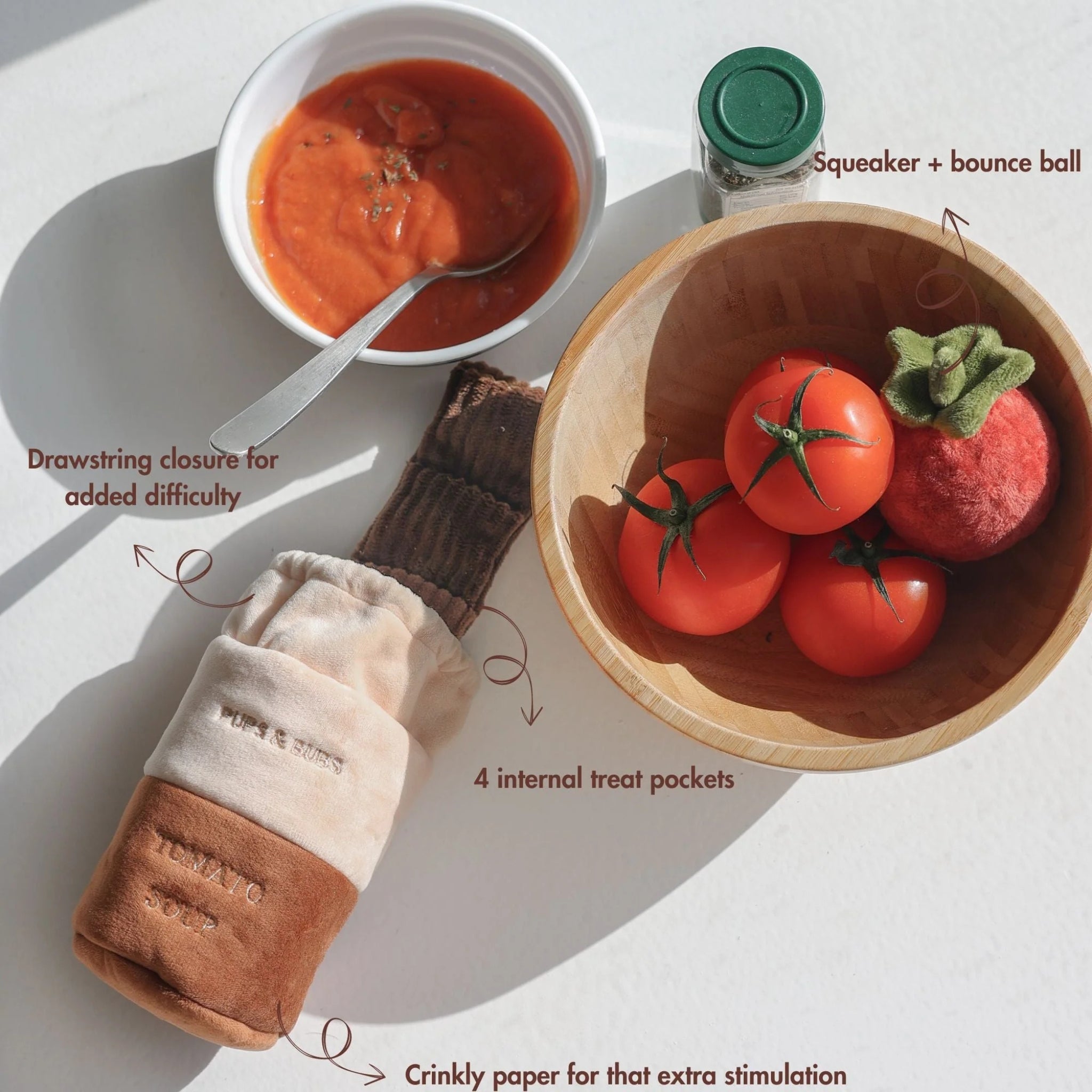 Top-down view of the tomato soup enrichment dog toy with labels pointing out the drawstring closure, internal treat pockets, crinkle paper, and squeaker ball.