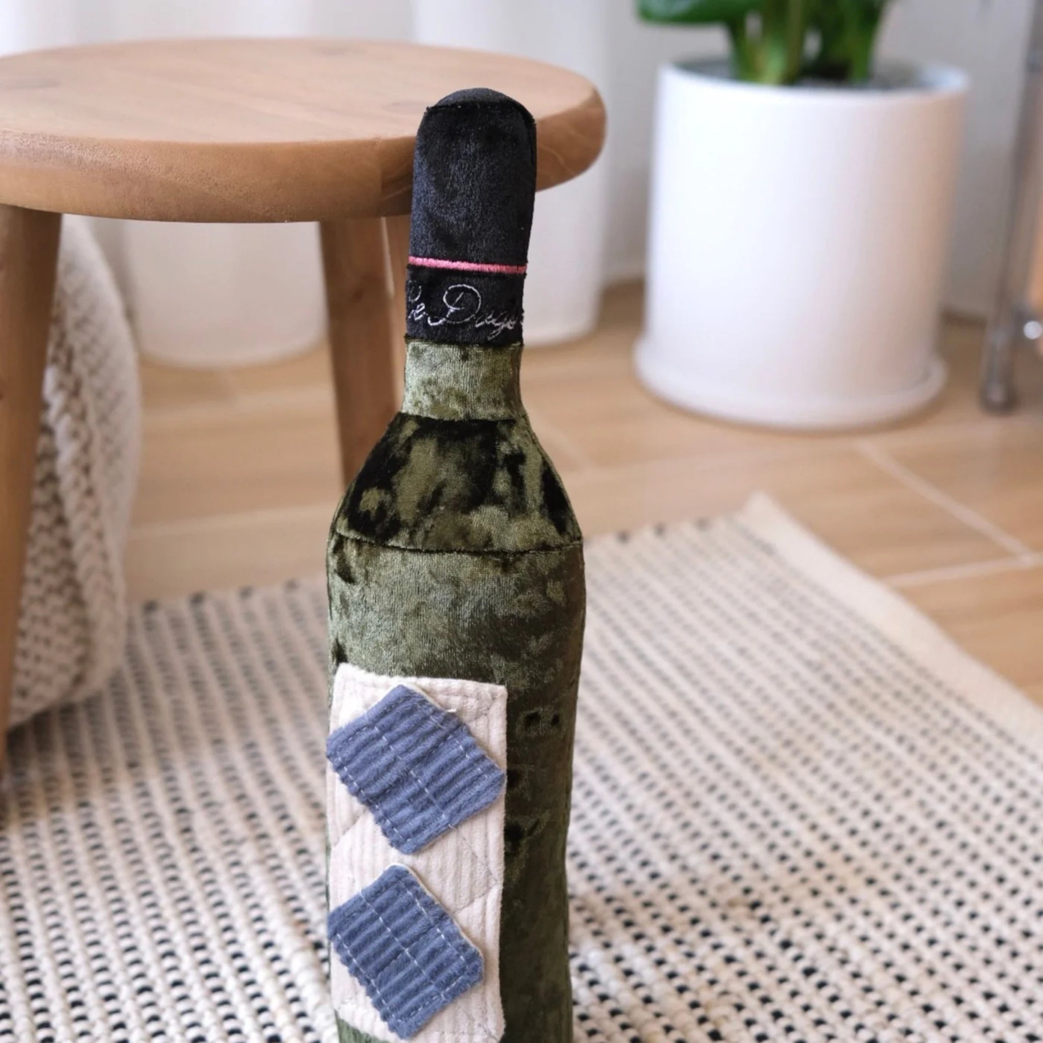 The velvet Merlot wine bottle dog toy standing upright on a patterned rug indoors next to a small wooden stool and a white potted plant.