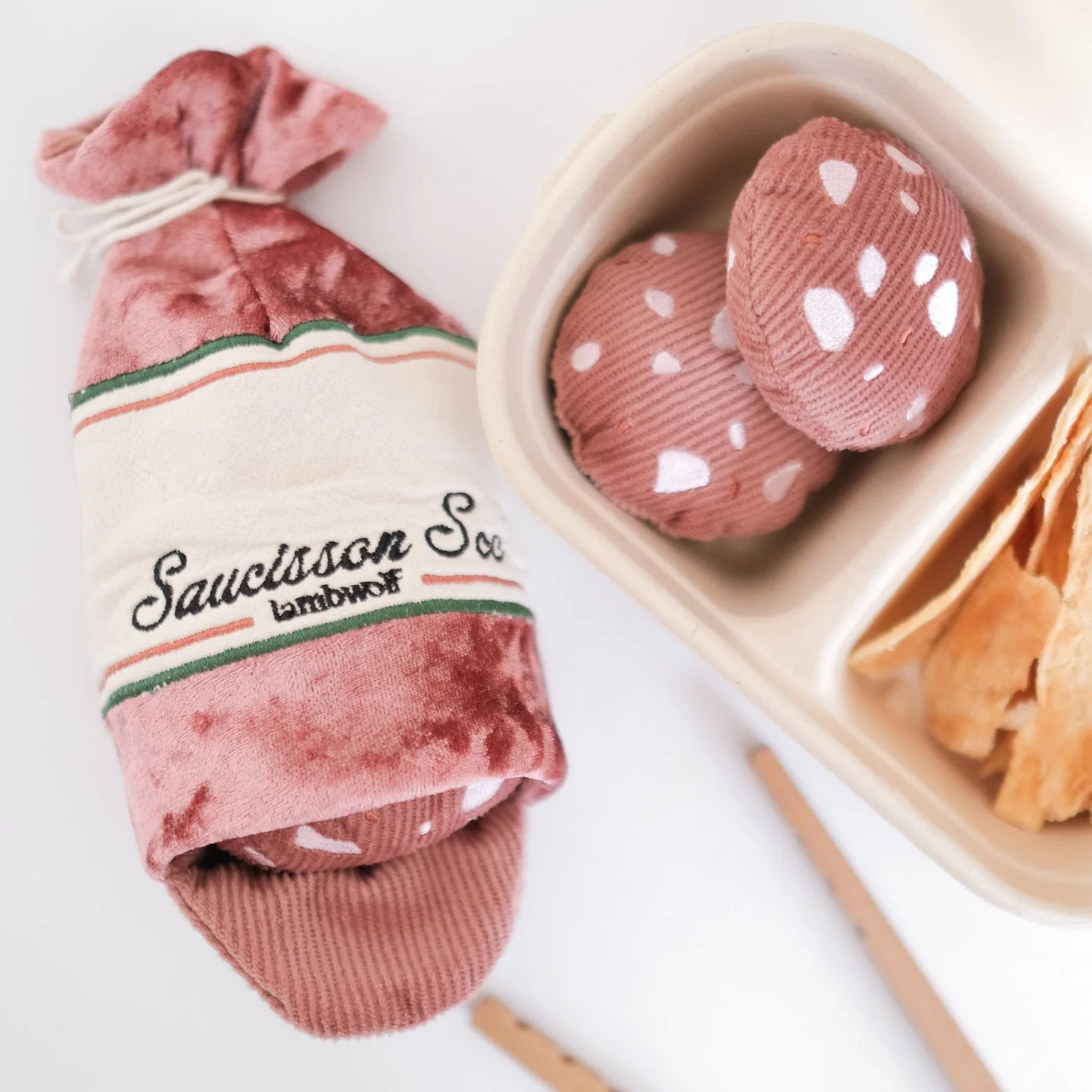 A lifestyle presentation of the interactive plush sausage dog toy next to a paper pulp bento lunch box filled with matching corduroy sausage slices and crunchy dog treats.