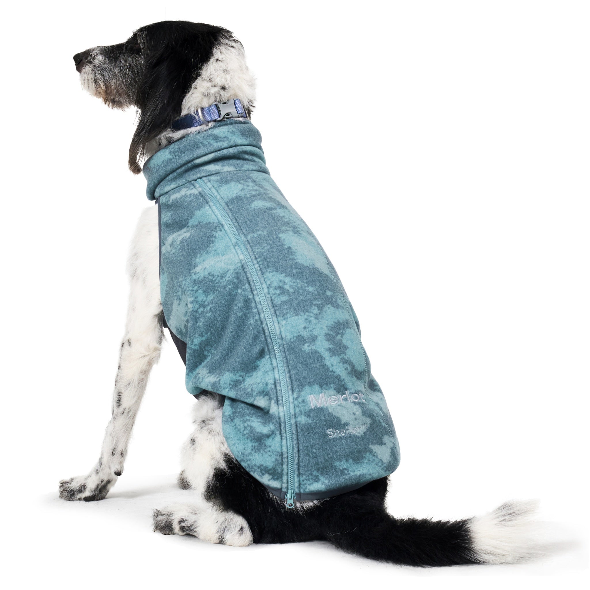 A three-quarter rear view of a sitting black and white dog, highlighting the ergonomic side zipper orientation and full hip coverage of the teal-patterned forest fleece pet coat.