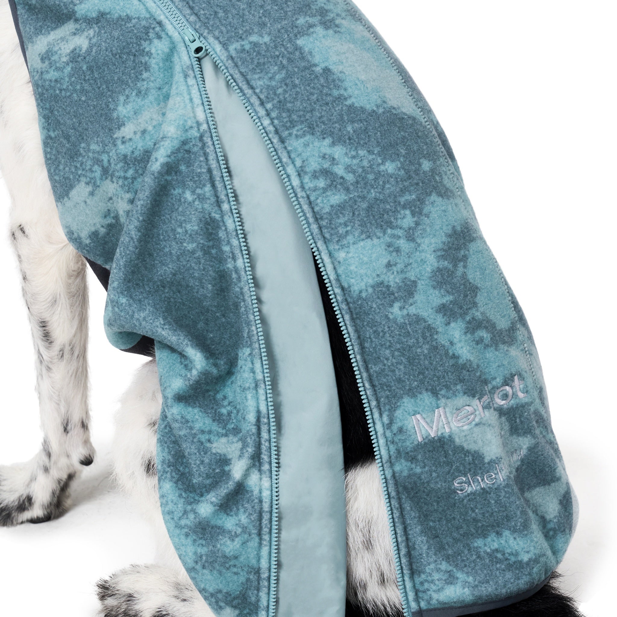 A detailed close-up shot focusing on the side zipper closure, the premium teal-green tie-dye fleece texture, and the silver embroidered Merlot brand name on the side of the dog jacket.