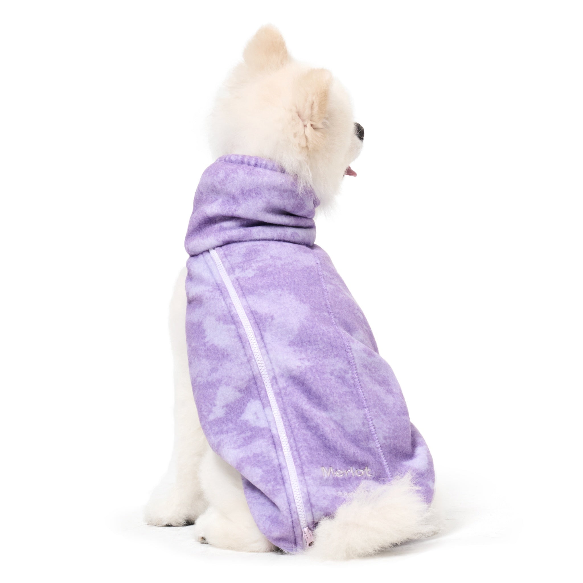 Rear view of a fluffy white small dog sitting down against a white background, wearing the purple tie-dye patterned fleece Merlot Shelter dog jacket featuring a white side zipper and an embroidered logo near the lower hem.