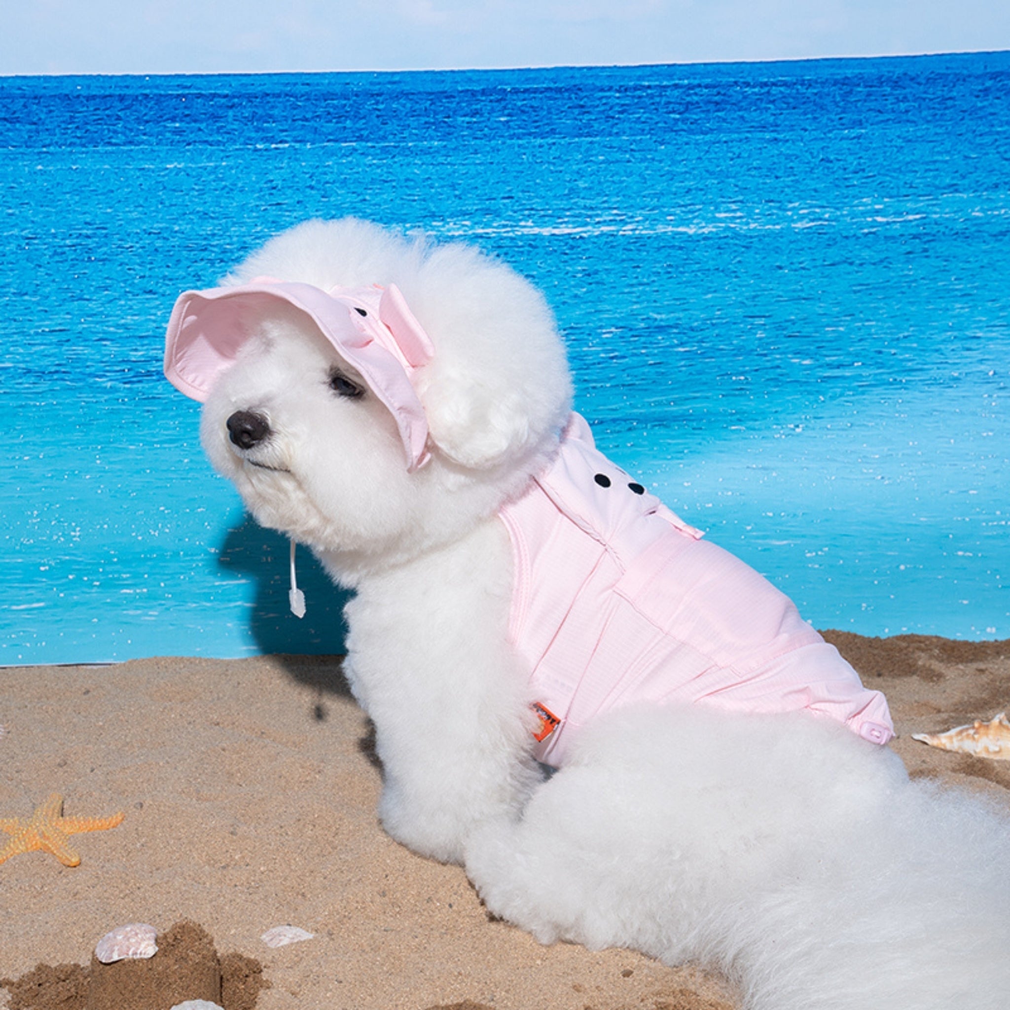 Side profile of a fluffy white Bichon Frise sitting on the sand against a bright blue ocean backdrop, wearing a matching baby pink wide-brim dog sun hat and cooling vest.