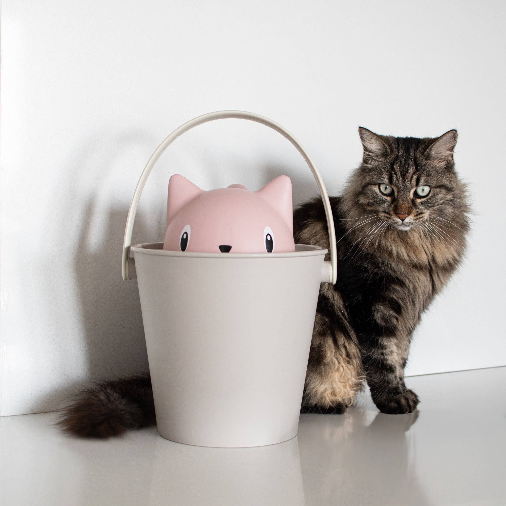 A lifestyle studio shot of a fluffy, long-haired tabby cat sitting next to the assembled Crick cat dry food container by Puppy Gallery, which features a taupe grey bucket with an upright handle and a glossy pink cat-shaped lid.