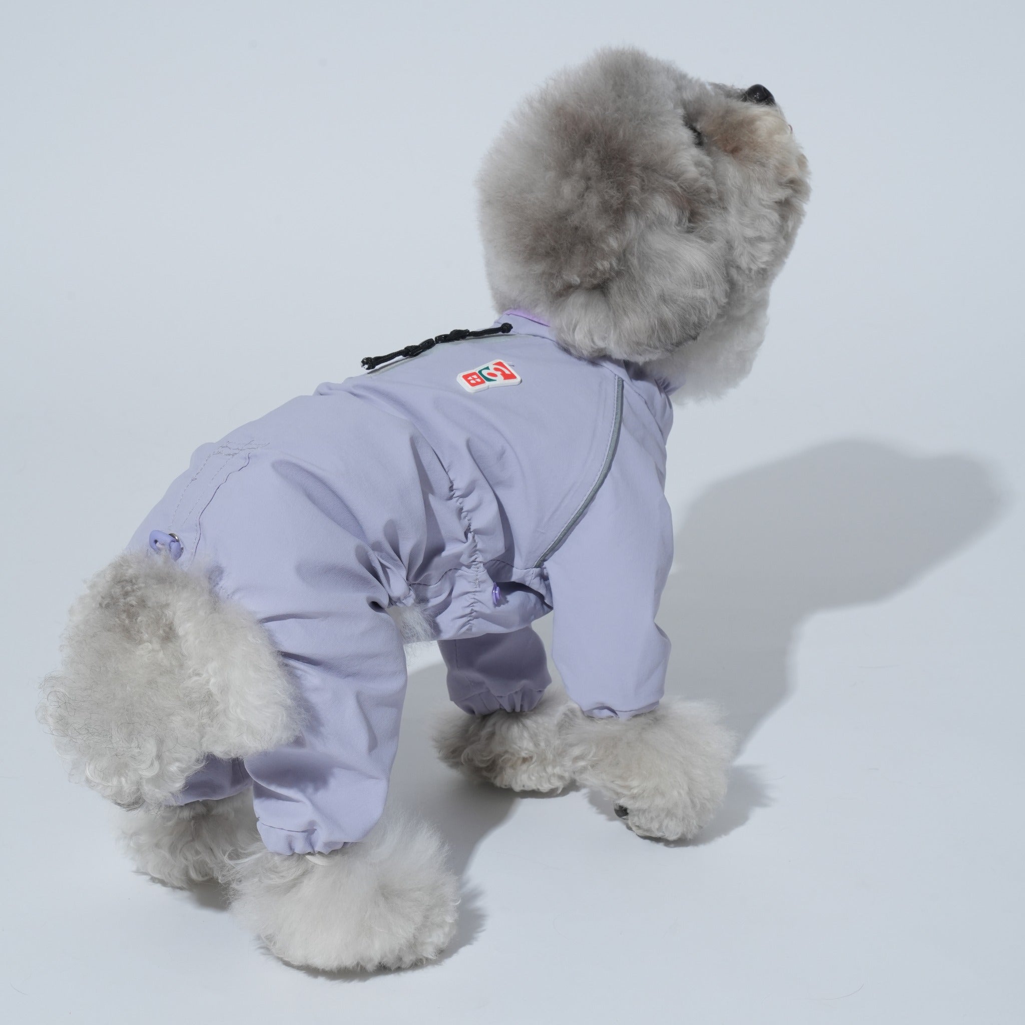 Rear side view of a grey poodle showcasing the adjustable drawstrings, reflective safety piping, and elasticized leg cuffs of the lavender Puppy Gallery rain jumpsuit.