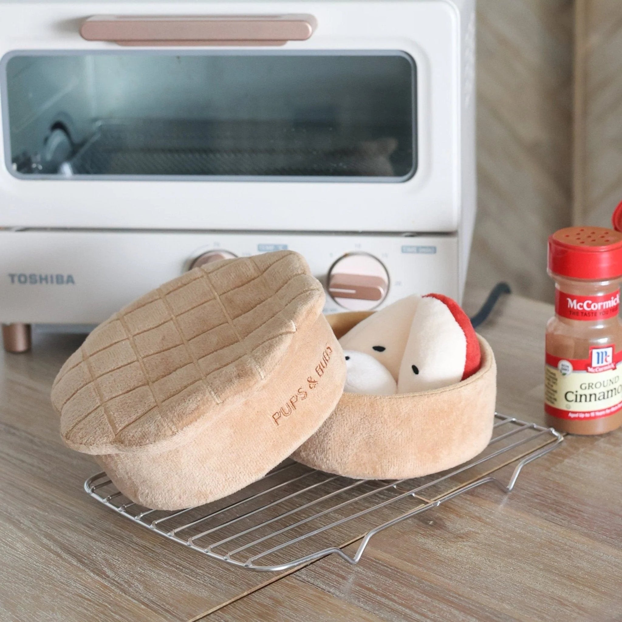 Plush interactive apple pie dog puzzle toy on a cooling rack next to a bottle of ground cinnamon in a kitchen setting.