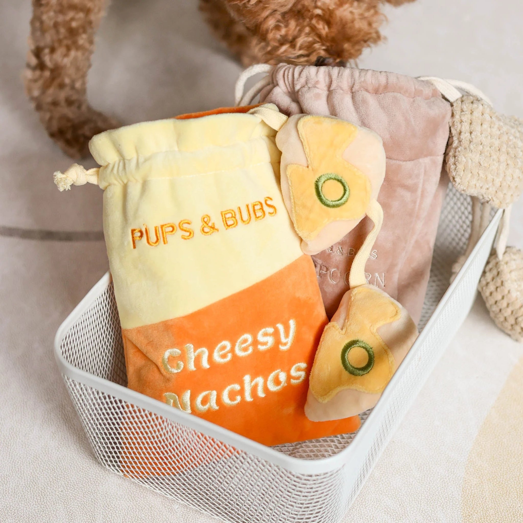 Cheesy Nachos and Popcorn plush nosework dog toys by Pups and Bubs neatly arranged in a modern white wire basket.