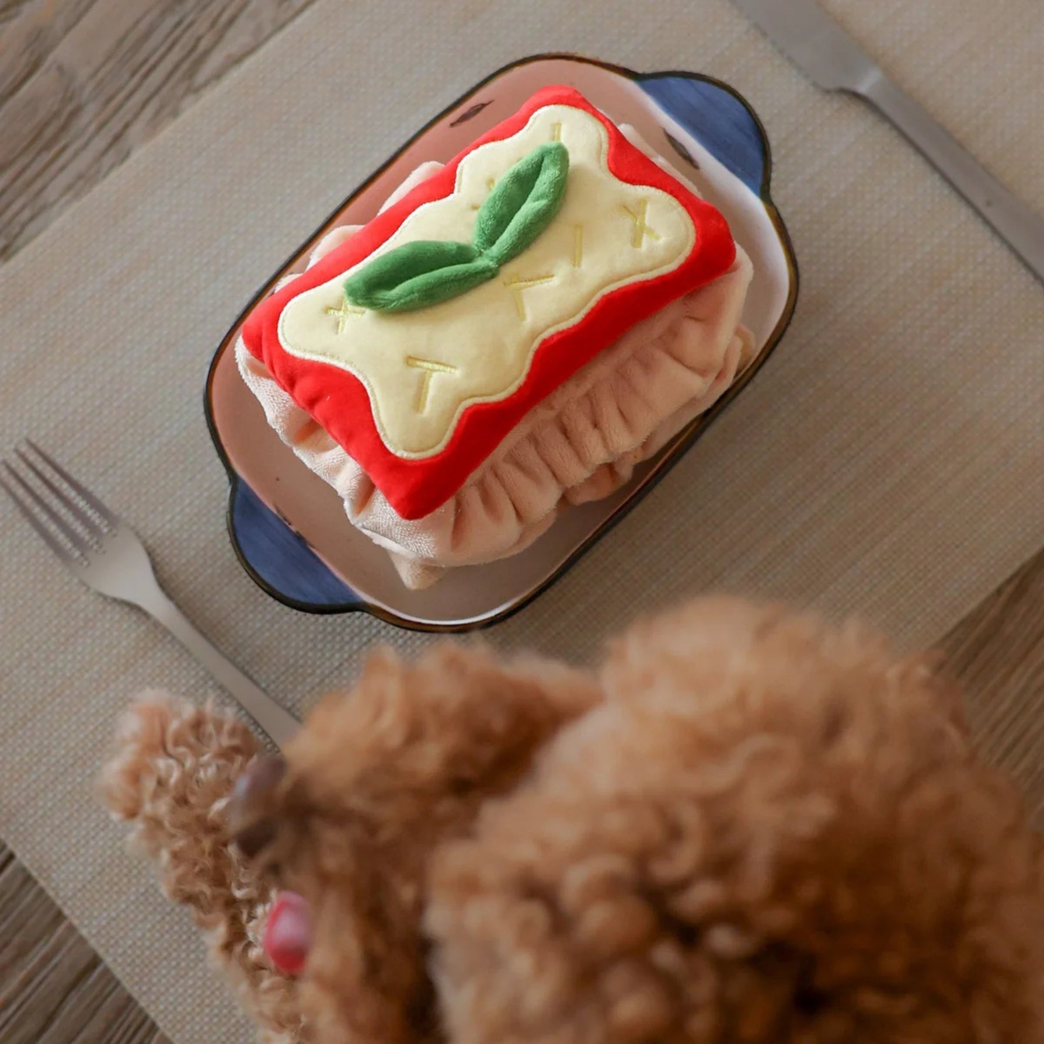 Overhead view of the Pups and Bubs Lasagna interactive plush dog toy served in a small baking dish on a table mat, with a blurry brown poodle looking on.