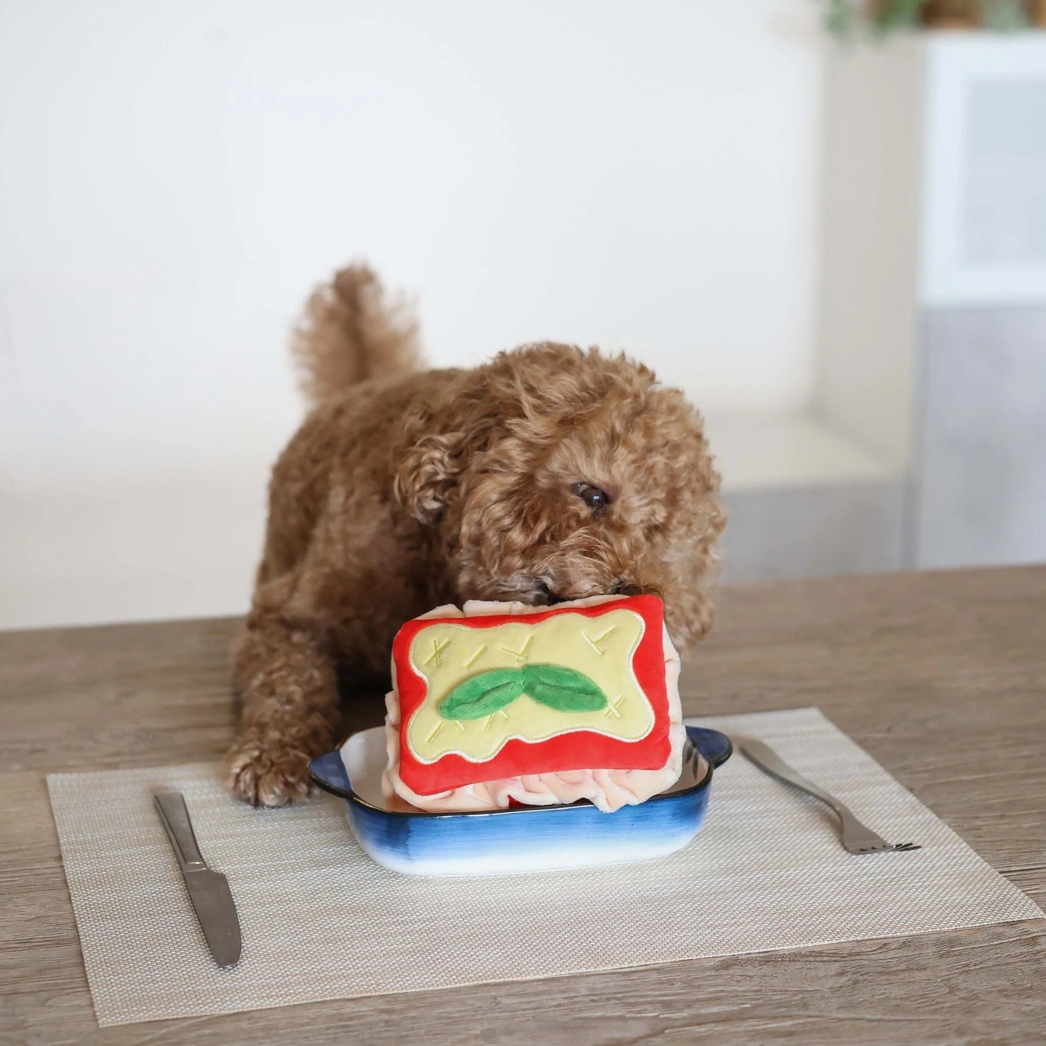 A brown poodle sitting at a wooden table playfully nudging the Lasagna interactive plush toy inside its small baking dish.