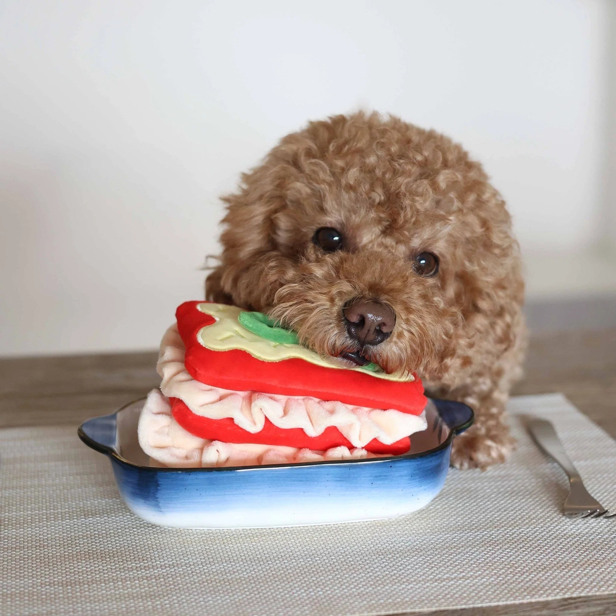 A fluffy brown poodle biting the side of the Lasagna nosework toy, which is layered with ruffled plush fabric and served in a blue and white dish.