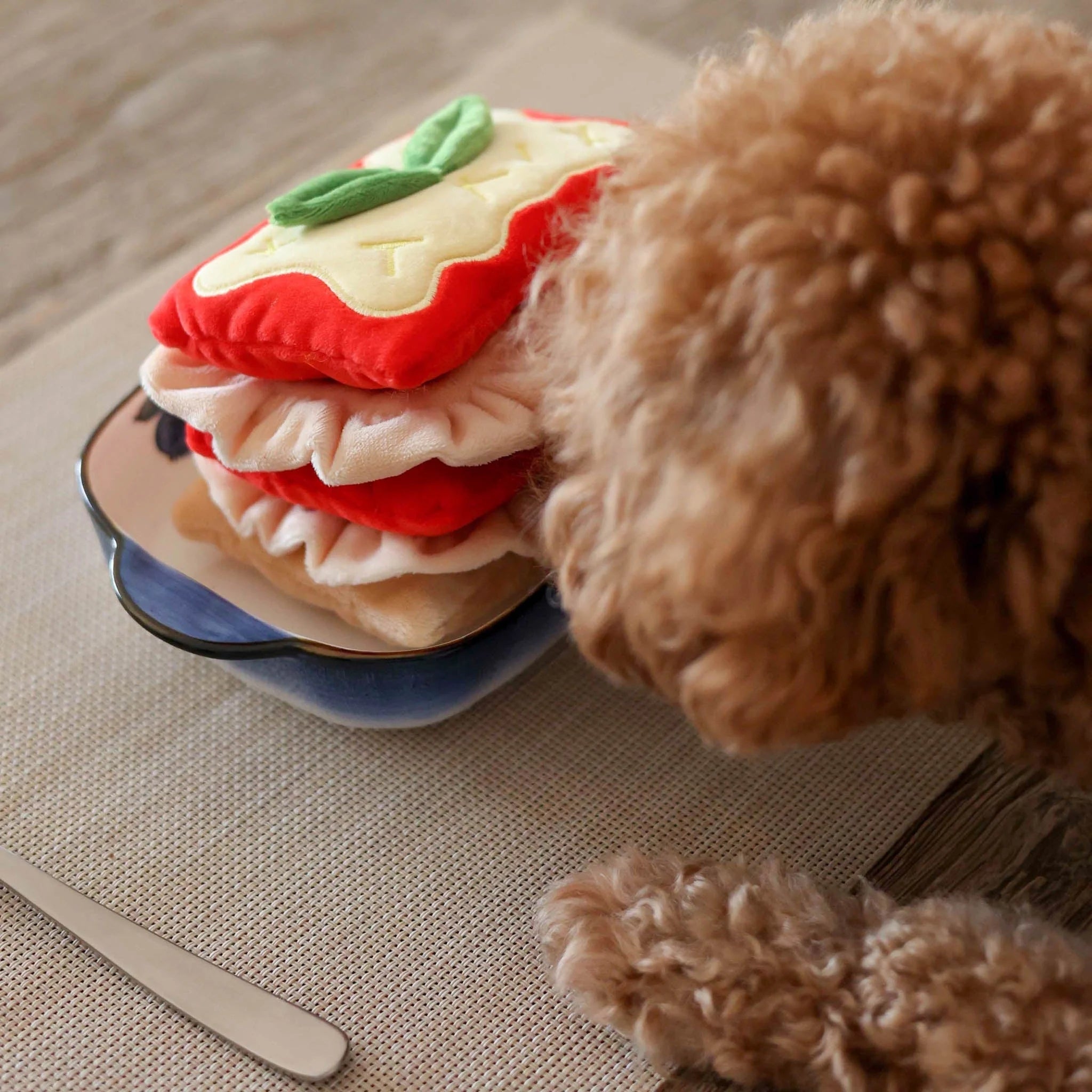 Side view of a curly brown poodle sniffing the edge of the multi-layered Lasagna plush dog toy to search for hidden kibble.