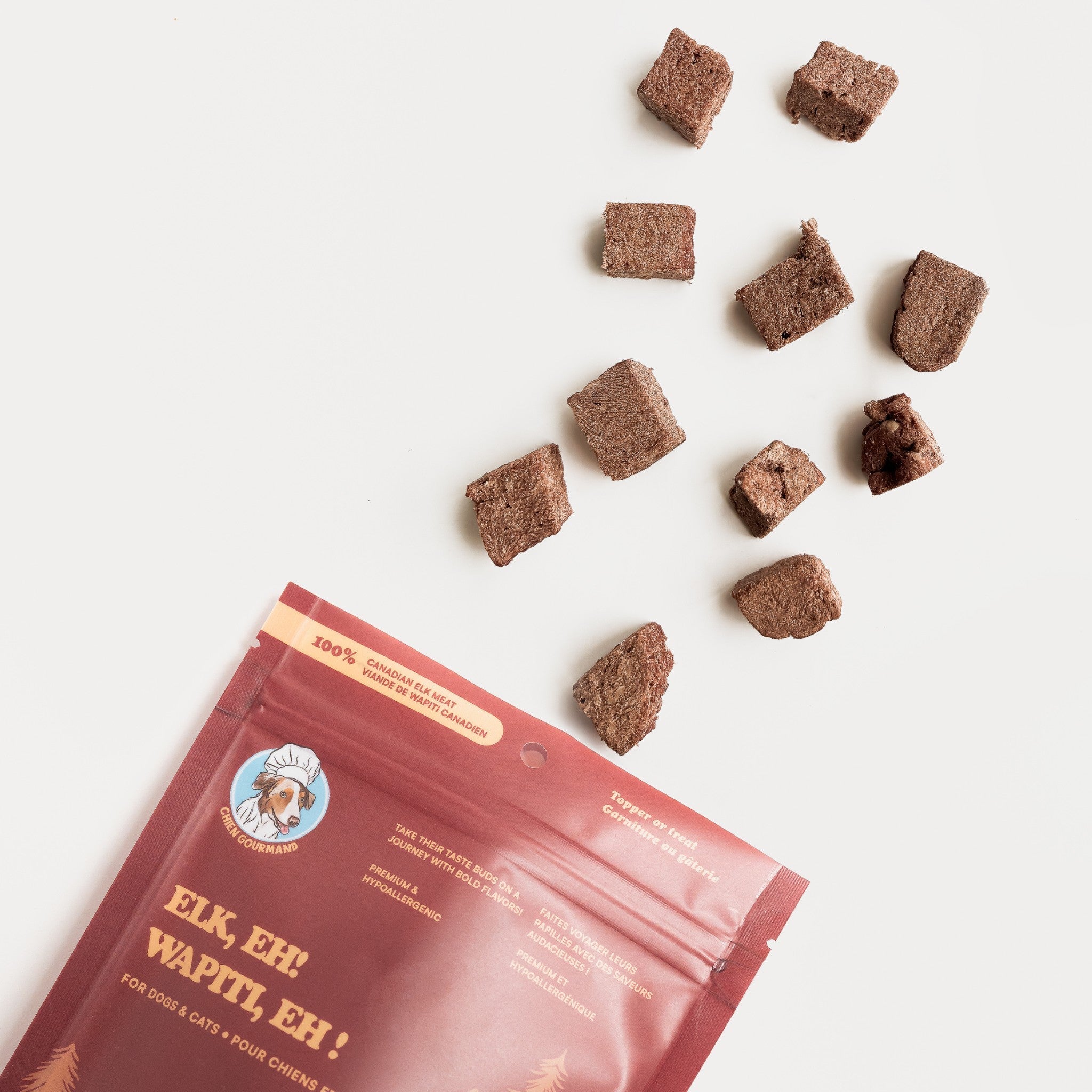 A top-down view of Chien Gourmand freeze-dried elk meat pieces tumbling out from a dark red product pouch onto a clean white surface.