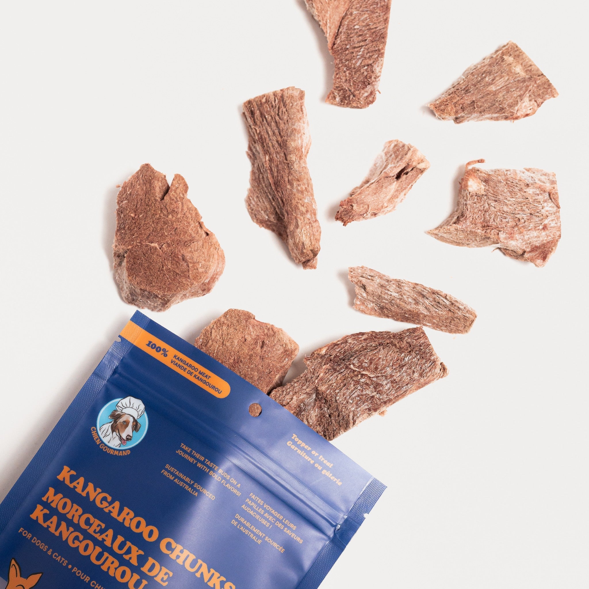 A top-down lifestyle view showing freeze-dried kangaroo meat chunks tumbling out of a blue Chien Gourmand product pouch onto a clean white surface.