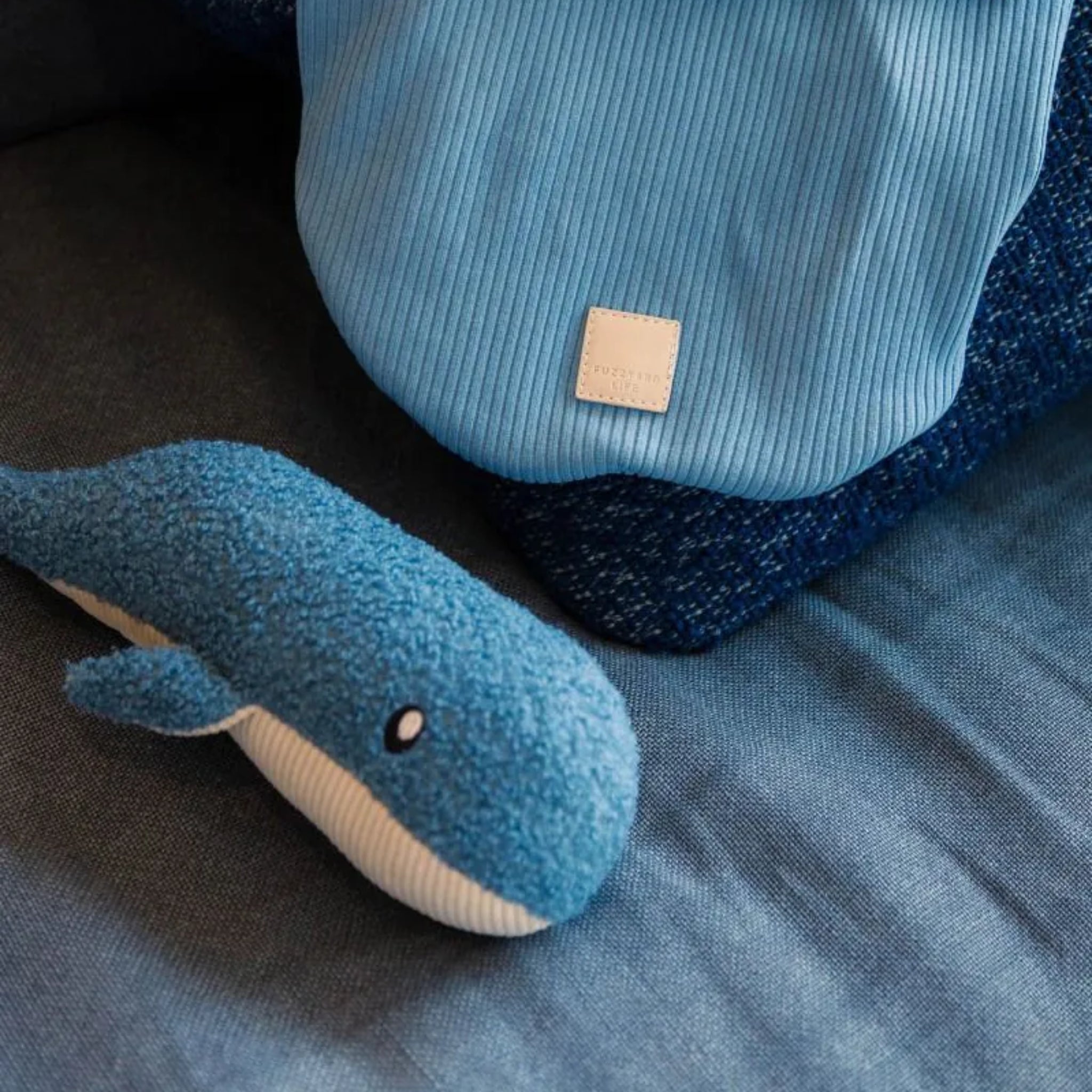 A cozy home lifestyle shot featuring the French blue whale plush dog toy resting on a dark grey sofa next to a matching light blue ribbed fabric cushion with a small square leather FuzzYard Life brand patch.