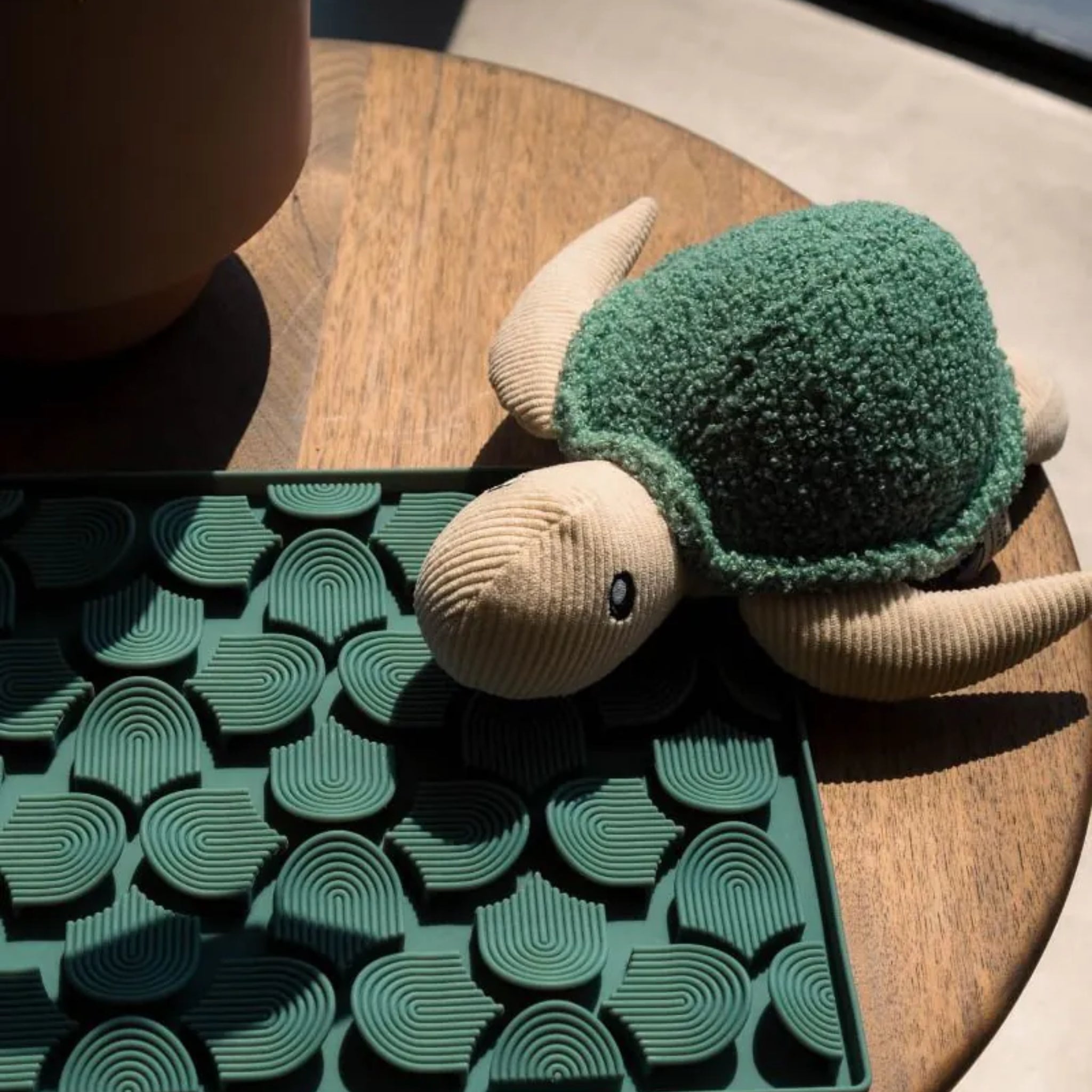 A sunlit lifestyle shot of the FuzzYard Life Myrtle Green Turtle plush dog toy resting on a round wooden table, positioned next to a deep green textured silicone pet licking mat.