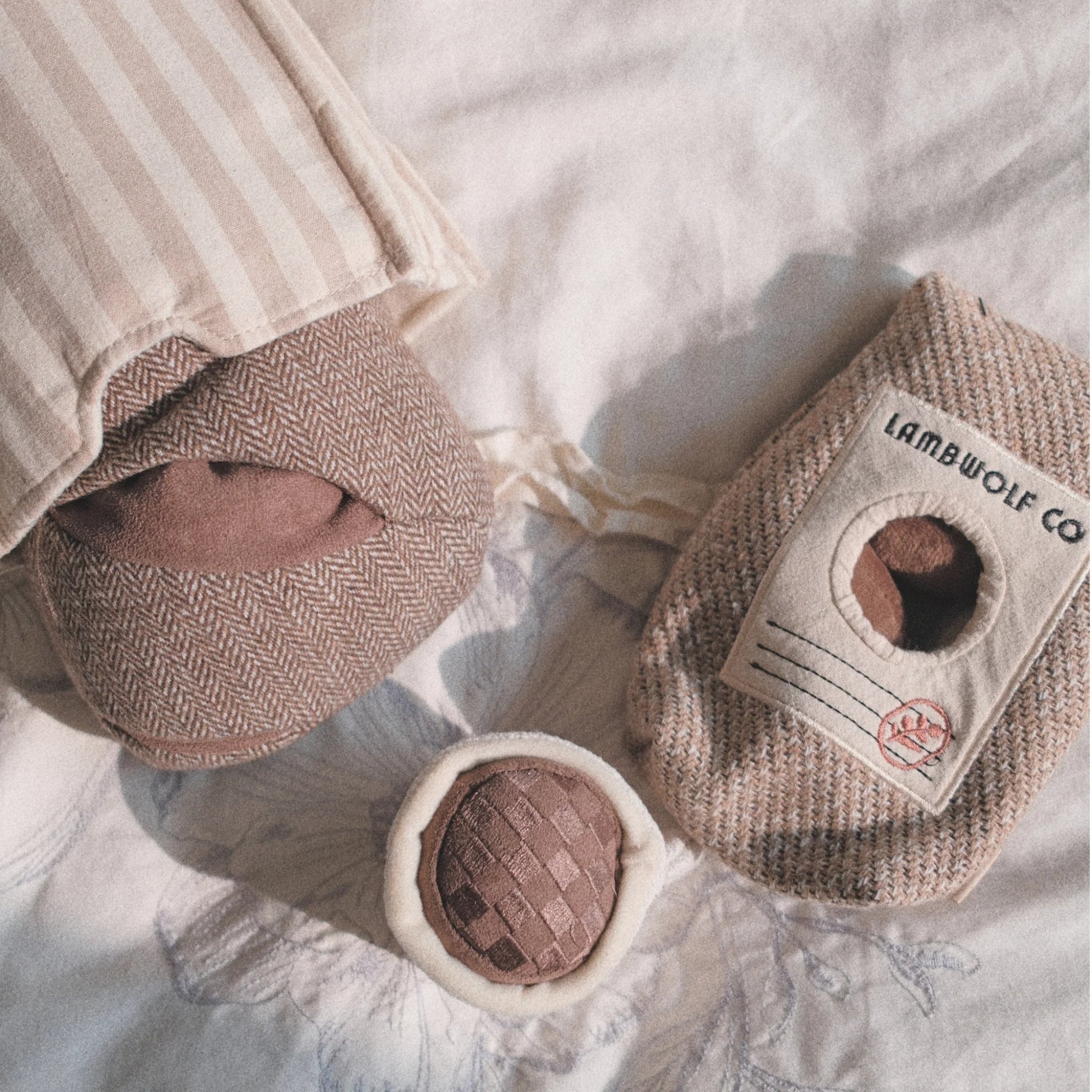 An overhead cozy aesthetic lifestyle flatlay of the coffee sack pet enrichment toy alongside matching neutral-toned plush herringbone treats on soft bedding.