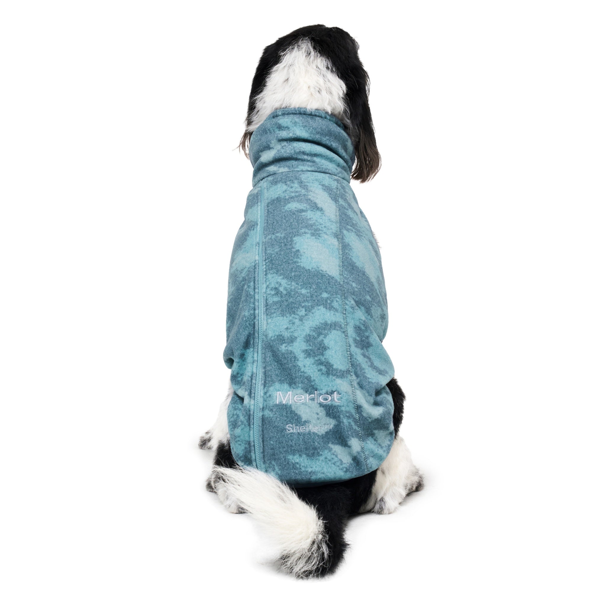 A direct back view of a sitting black and white dog facing away from the camera, highlighting the clean spine stitching and subtle silver Merlot embroidery on the lower hem of the forest green fleece coat.