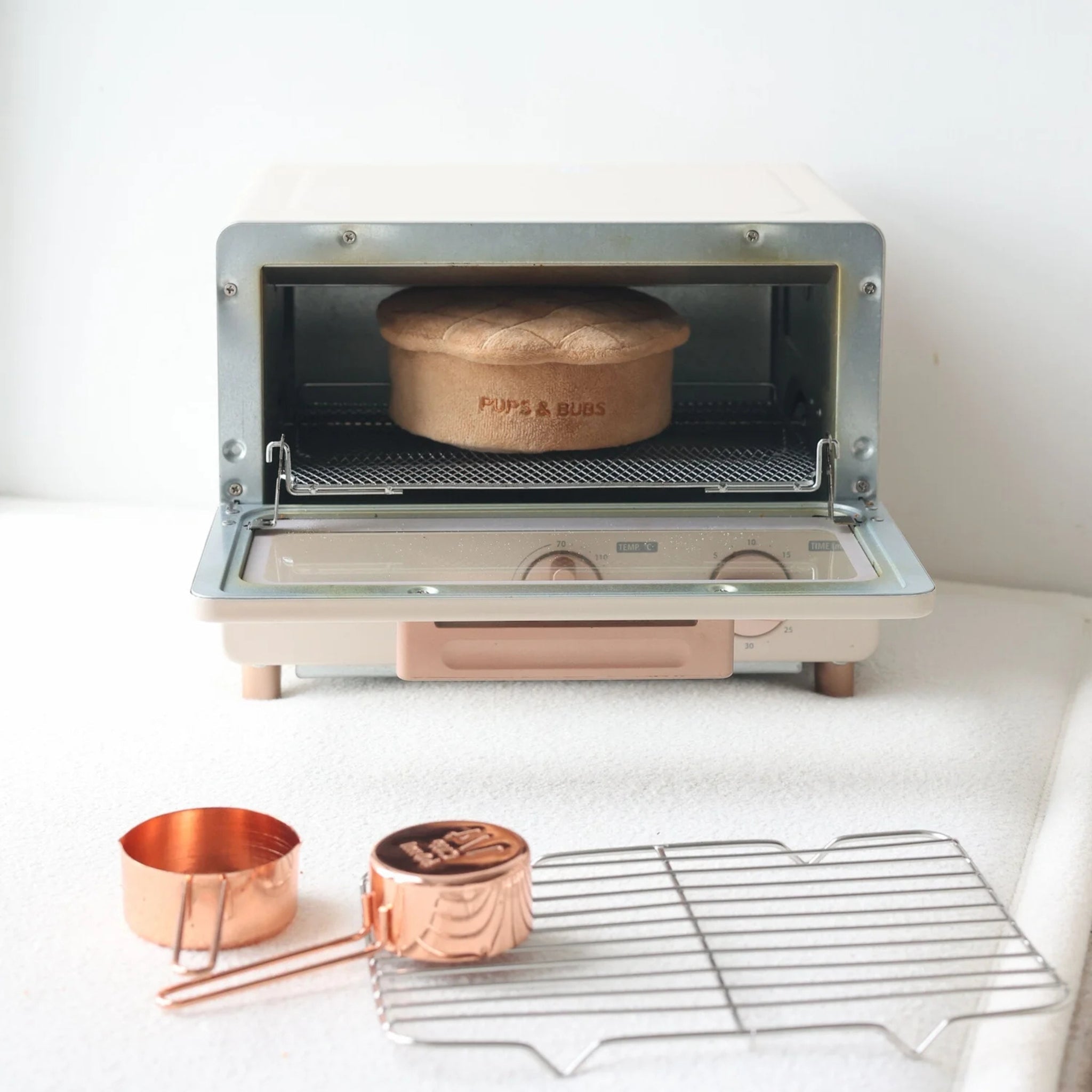 Plush apple pie nosework dog toy by Pups and Bubs sitting inside a cute miniature white toaster oven on a baking mat with measuring cups